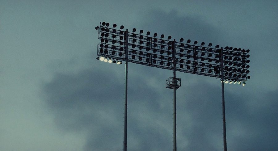 Low angle on a stadium light bank at dusk, three stanchions, no ground and no horizon, against a flat blue grey sky