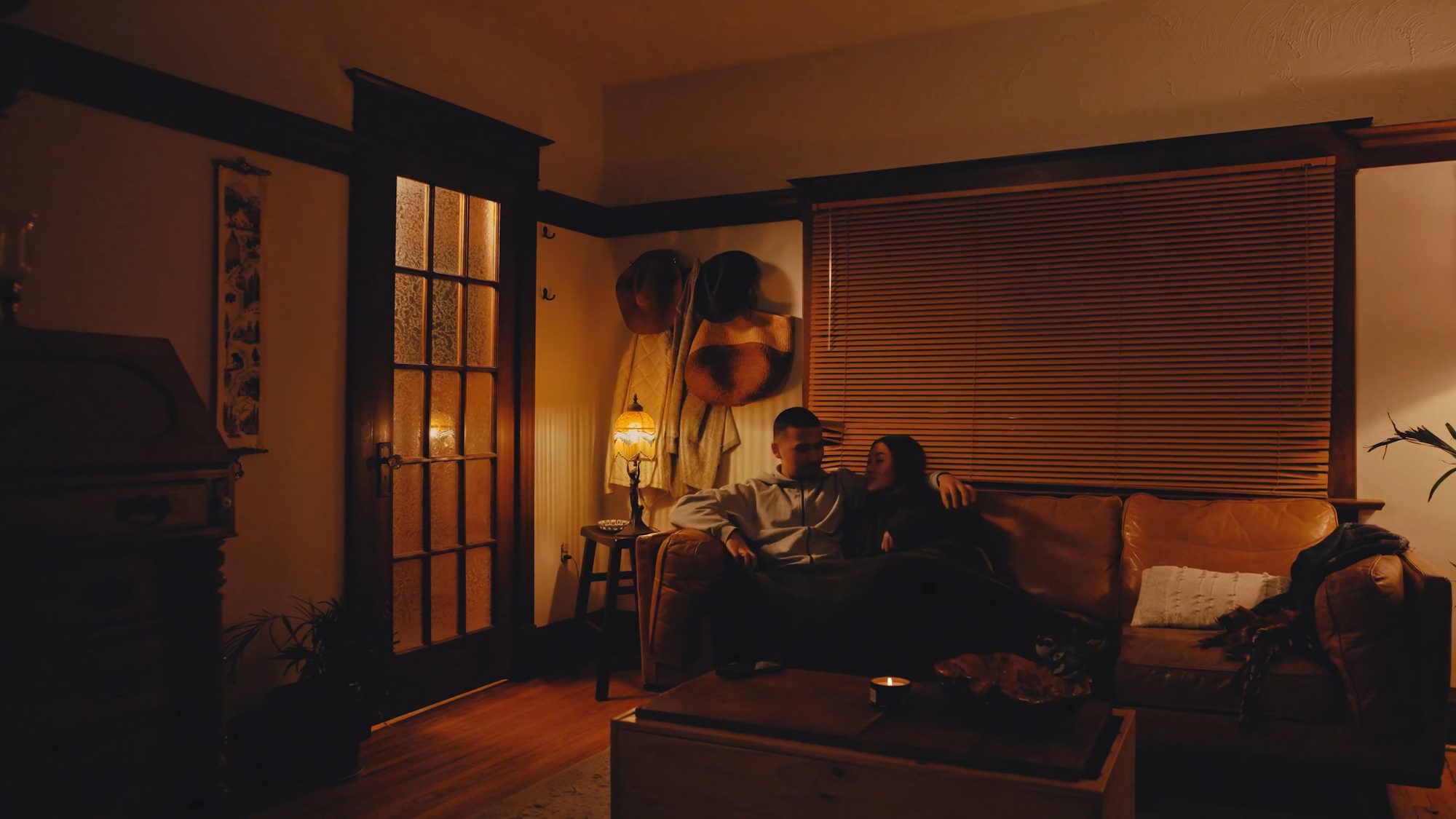 A front room at night, a couple settled together on a leather couch under a stained glass table lamp, wood trimmed french doors and drawn blinds around them