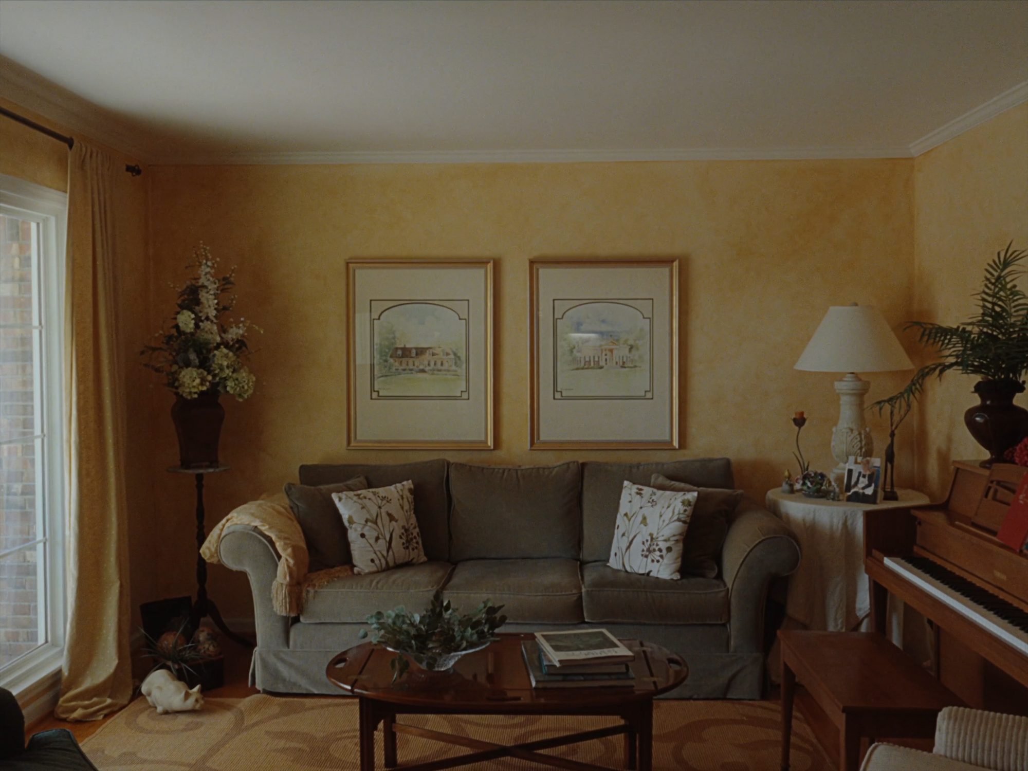A warm living room in window light, framed pictures over the couch, an upright piano and a lamp, no one in the room