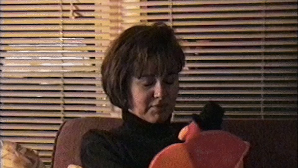 Warm four by three home video interior, a woman seated in front of venetian blinds, heavy film grain