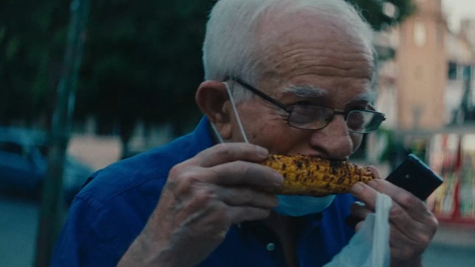 Blue hour close portrait of an older man in glasses eating elote on a street, his face exposed and the background fallen away to nothing