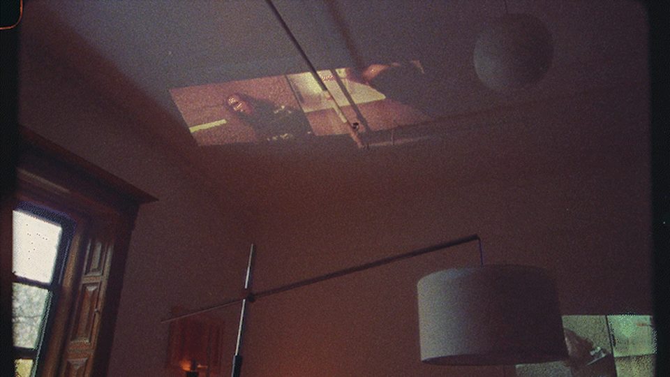 A dim living room at dusk, a lamp on at the left, television light thrown across the ceiling, the source out of frame