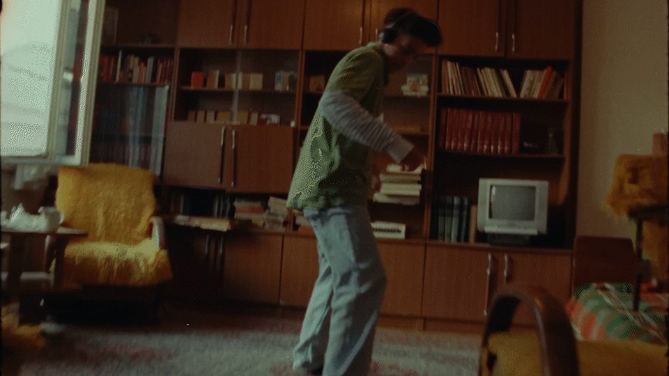 A figure dancing alone in a wood panelled front room with a wall unit, books, a boxy television and a yellow armchair