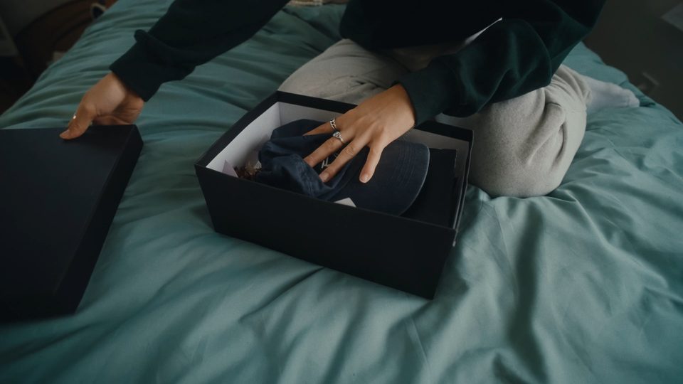Low light teal bedroom, two hands opening a box on a bed with a cap folded inside